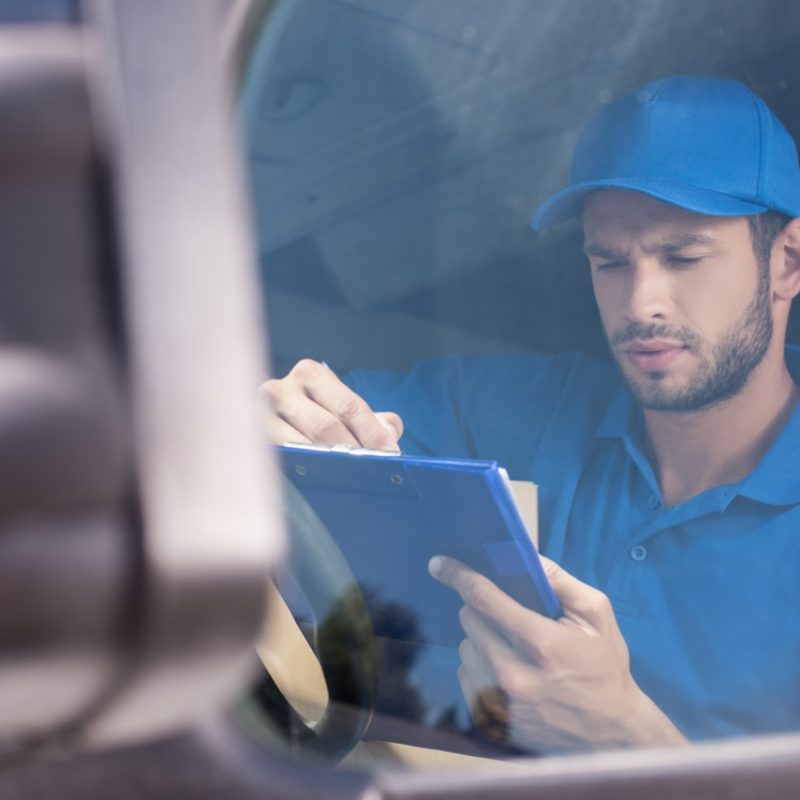handsome delivery man filling in documents on clipboard at car